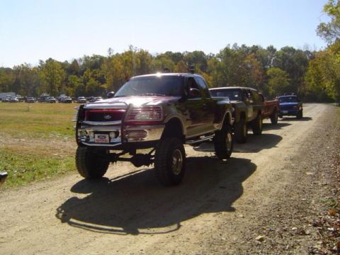 1979 Ford Bronco - A line of modified trucks drives down a dirt road on a sunny day with trees in the background.