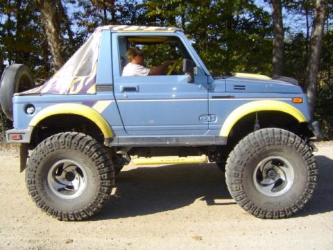 1979 Ford Bronco - A blue Suzuki Samurai with off-road tires is parked on a dirt road with trees in the background.