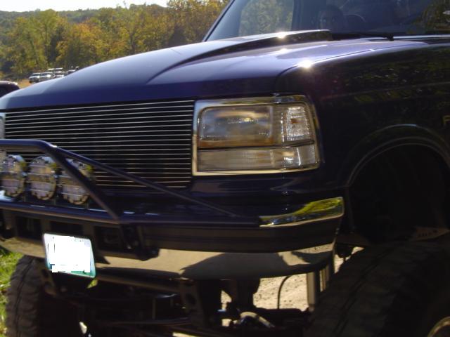 Front end of a dark blue Ford F-Series truck with a chrome grille and aftermarket bumper with lights. License plate is obscured