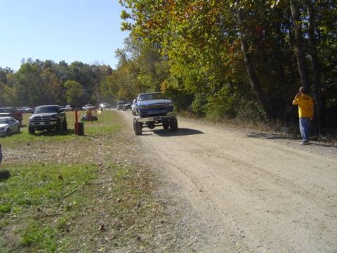 1979 Ford Bronco - A blue pickup truck is transported on a trailer along a dirt road, with several other vehicles visible in the background.