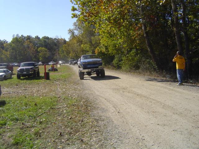 Blue pickup truck being towed on a trailer down a dirt road. Other trucks and cars parked on the side