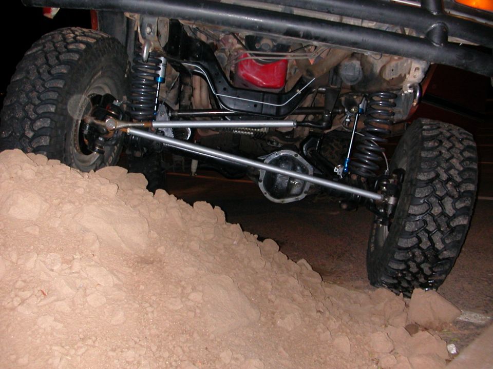 Off-road vehicle's rear axle, suspension, and tires on a dirt mound. Visible components include shocks, springs, and differential