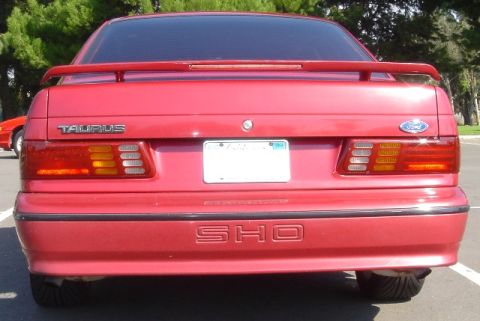 1991 Ford Taurus - Straight-on rear view of a red first-generation Ford Taurus SHO sedan parked in a lot, showing the raised trunk-lid spoiler, TAURUS and For…