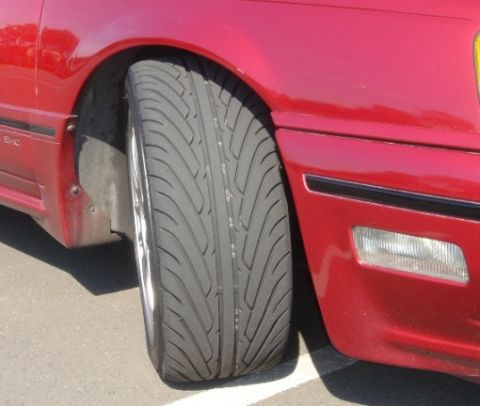 1991 Ford Taurus - Close-up of a red Ford Taurus SHO's left front corner with the wheel removed and a wide directional-tread tire on a dark wheel propped agai…