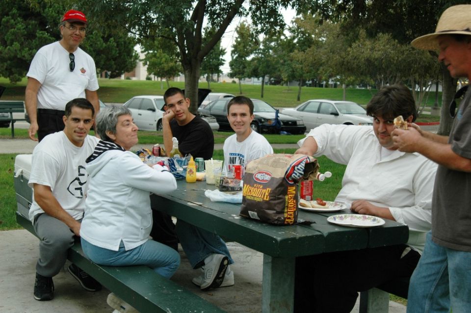 People gathered around a picnic table with cars parked in the background