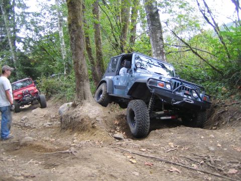 1990 Ford Bronco - Two off-road vehicles navigate a steep, wooded trail with one vehicle climbing over an obstacle.