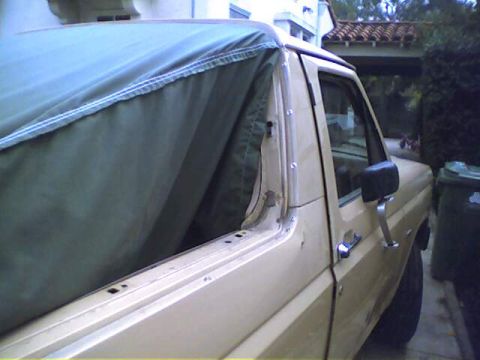 1994 Ford Bronco - A tan Ford Bronco is partially covered by a green soft top, showing its side profile and door.