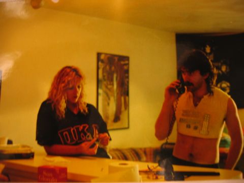 1985 Ford LTD Crown Victoria - A woman with blonde curly hair looks down at something on a table while a man with a mustache talks on a corded phone.