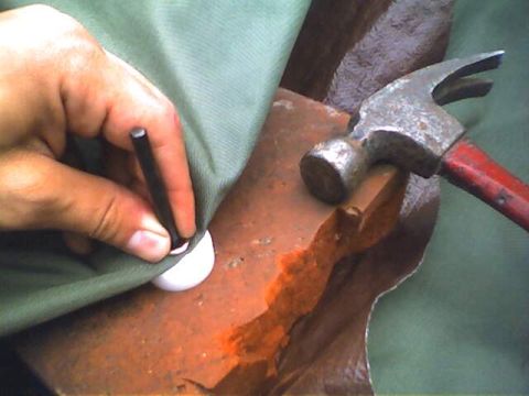 1994 Ford Bronco - A person is using a punch tool and hammer to install a grommet into a piece of green fabric.