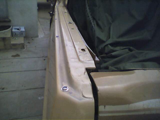 Close-up of a tan car body panel, possibly a fender or door, with a dark green tarp covering part of it. Some bolts are visible along the panel's edge