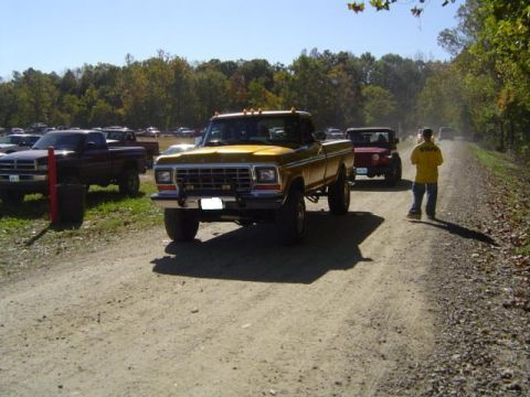 1979 Ford Bronco - A yellow 1979 Ford pickup truck is seen from the front on a dusty dirt road, with other vehicles and trees in the background.