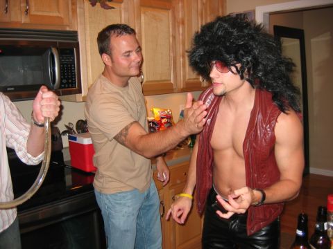 1985 Ford LTD Crown Victoria - A party scene in a kitchen with two men interacting, one dressed in a flamboyant costume.