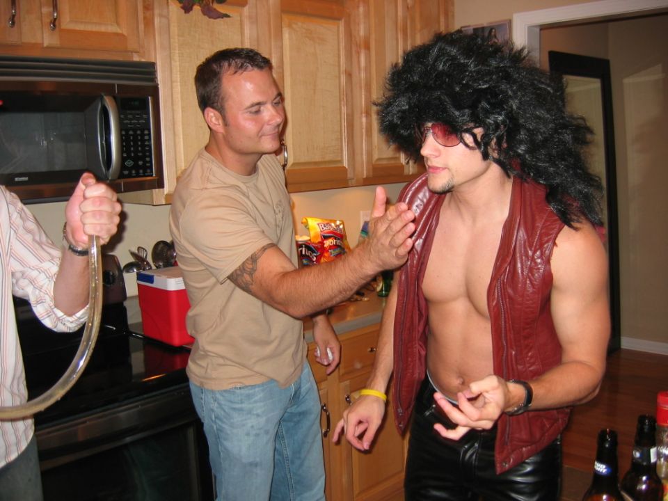 Two guys talking in a kitchen, one in a wig and sunglasses, the other holding a beer tube