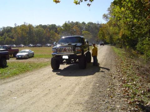 1979 Ford Bronco - A heavily modified black pickup truck sits on a dirt road, with parked vehicles and trees in the background.