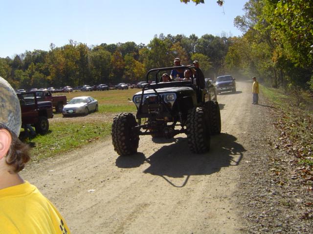 Off-road vehicle with large tires on a dirt road, people inside, trees and parked cars in background