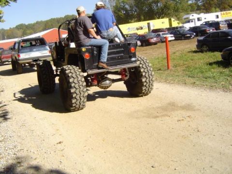 1979 Ford Bronco - Two people ride in a heavily modified off-road vehicle at an outdoor event with many other cars.