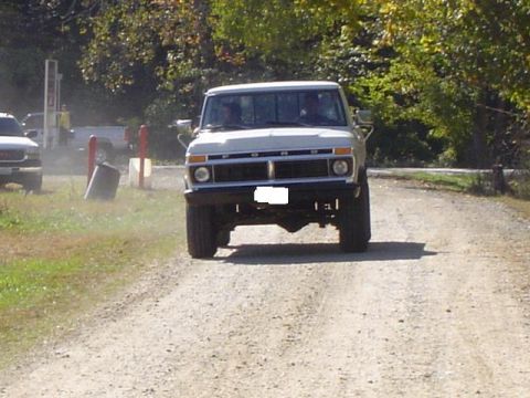 1979 Ford Bronco - A white pickup truck drives on a dirt road, with another vehicle visible in the background.