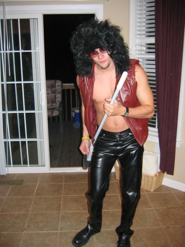 Man wearing a black wig, sunglasses, red vest, and black leather pants, holding a mop handle