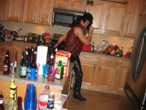 1985 Ford LTD Crown Victoria - A person in a leather outfit dances in a kitchen filled with party supplies and bottles.