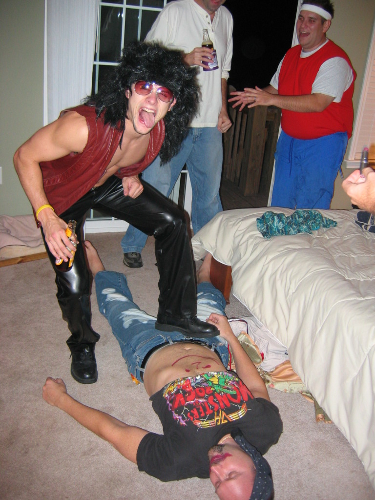 guy in wig and leather pants standing over another guy on the floor, two other guys watching