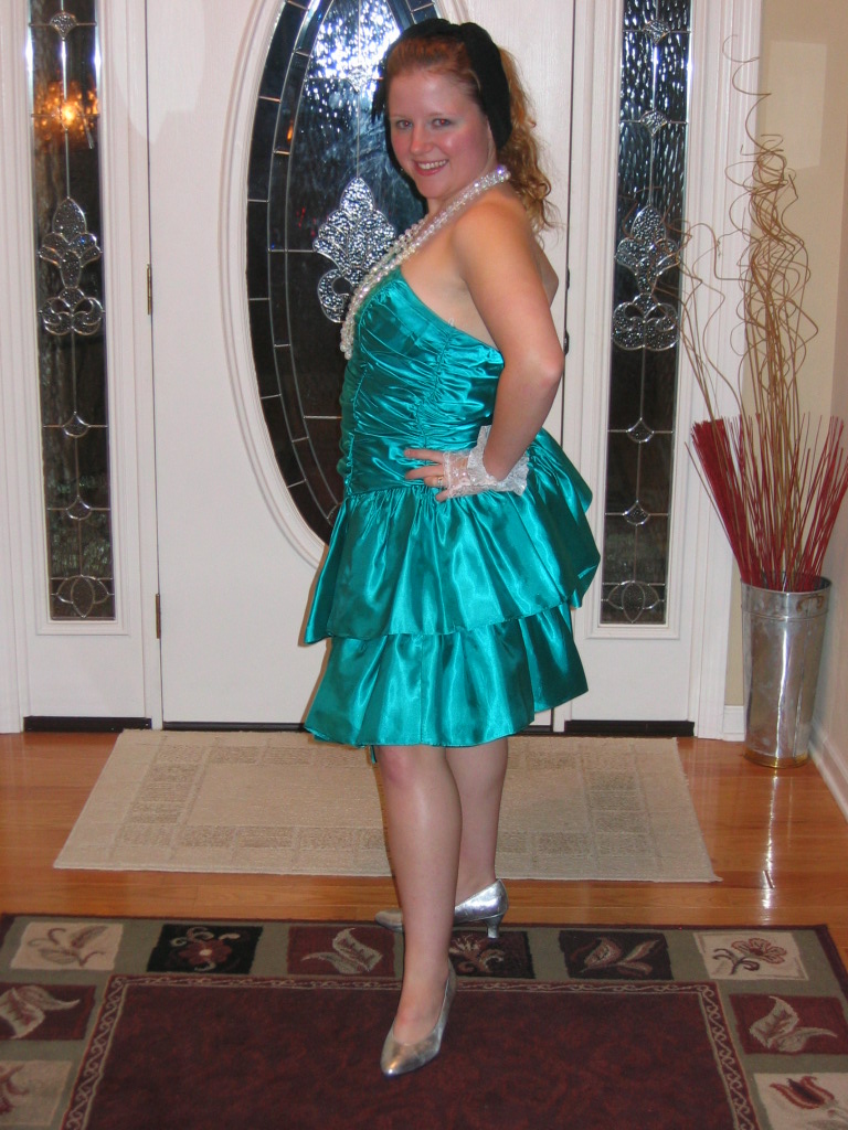 Woman in a teal, ruffled dress and pearls poses in front of a door