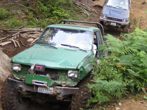 1990 Ford Bronco - Two off-road vehicles navigate a muddy, wooded trail with dense foliage.