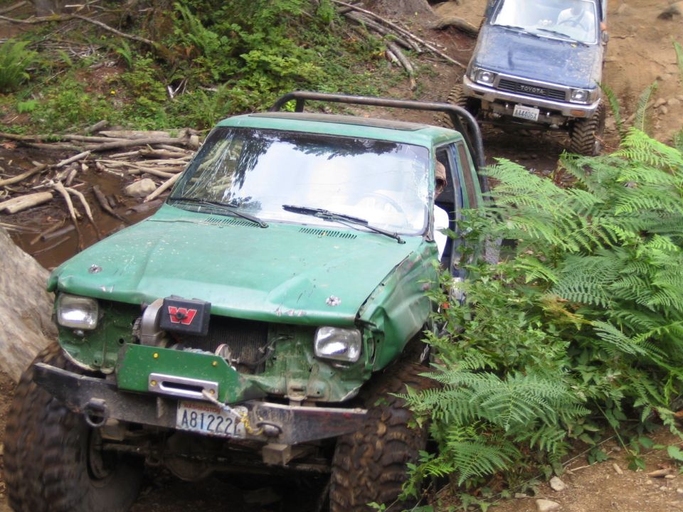 Green off-road truck with winch and damaged front end, blue Toyota in background on trail