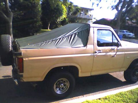 1994 Ford Bronco - A tan Ford Bronco from the 1980s is parked outdoors, covered with a dark green soft top and a spare tire on the back.