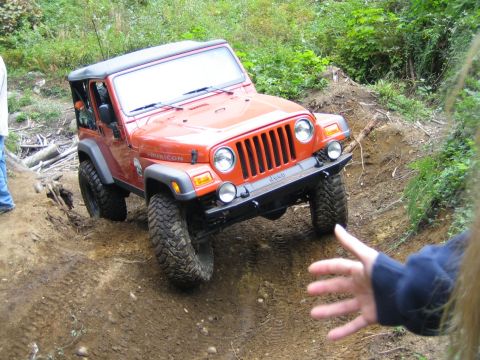 1990 Ford Bronco - An orange Jeep Wrangler Rubicon navigates a muddy off-road trail with trees in the background.