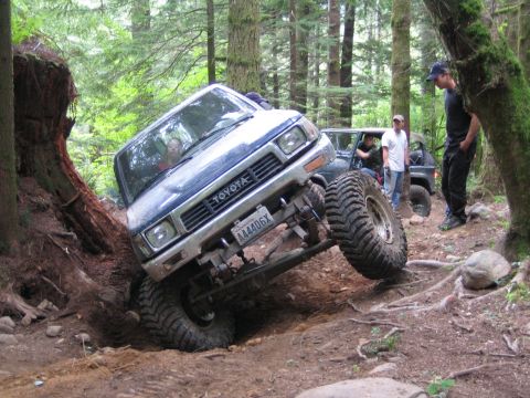 1990 Ford Bronco - A blue Toyota truck is shown attempting a difficult off-road obstacle with another vehicle visible in the background.