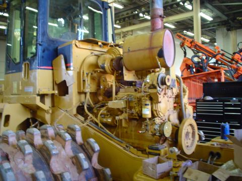 1966 International School Bus - Close-up view of the engine and drive train of a large yellow construction vehicle, with another piece of equipment visible behind it.
