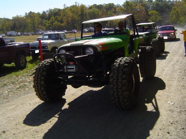 Green off-road buggy with large tires and a white roof, driving on a dirt road. Two people are in the front seats. Another similar vehicle follows behind