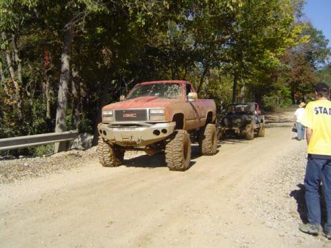 1979 Ford Bronco - A lifted red GMC pickup truck leads another off-road vehicle down a dusty path during an event.