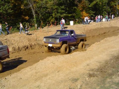 1979 Ford Bronco - A purple pickup truck navigates a muddy track during an off-road event with spectators in the background.