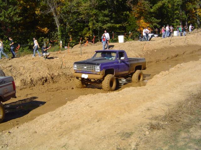 Purple pickup truck with lifted suspension and large tires driving through mud, front grille visible