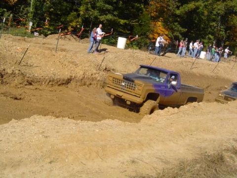1979 Ford Bronco - A purple and brown pickup truck navigates a muddy off-road course with onlookers in the background.
