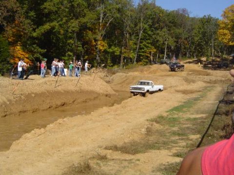 1979 Ford Bronco - Several trucks navigate a muddy off-road course with onlookers watching from the embankment.