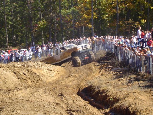 White off-road truck with large tires climbing a dirt hill, crowd watching behind a fence