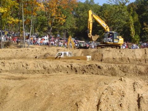 1979 Ford Bronco - A tan pickup truck is stuck in a mud pit during an event, with an excavator nearby and spectators watching.