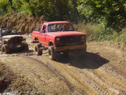 1979 Ford Bronco - Two off-road vehicles navigate a wet, muddy path through a wooded area.