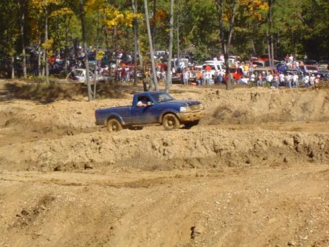 1979 Ford Bronco - A blue pickup truck navigates a muddy course during an event with many vehicles and people watching.