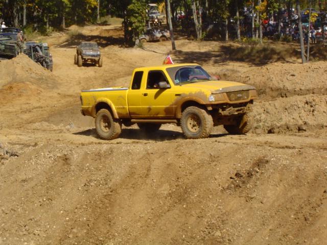 Yellow pickup truck driving on a dirt track with other vehicles and spectators in the background