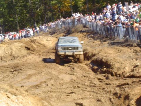 1979 Ford Bronco - A blue pickup truck navigates a muddy track during an event, with a crowd observing from above.