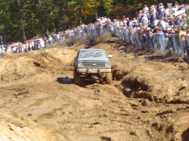 Blue pickup truck driving through mud, front view. Spectators watch from a raised embankment