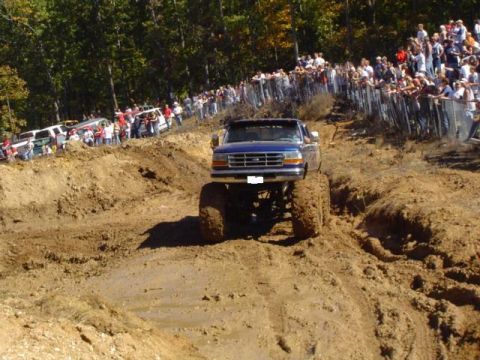 1979 Ford Bronco - A lifted blue pickup truck navigates a muddy track during an event, with spectators lined up on a hill.