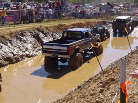 1979 Ford Bronco - A lifted pickup truck navigates a muddy course during an event, with other vehicles and spectators in the background.