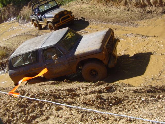 Muddy dark SUV stuck in mud, orange strap attached to front, another Jeep behind it