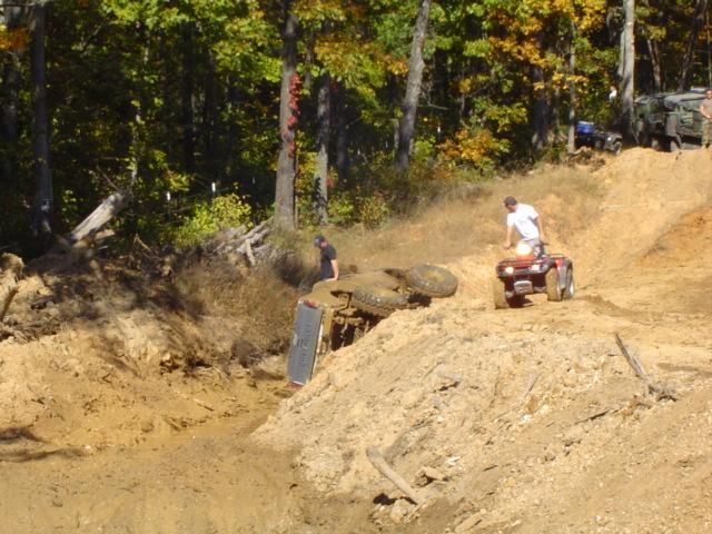 Overturned vehicle on its side in mud, with ATVs nearby on a dirt hill
