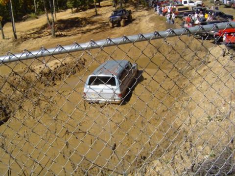 1979 Ford Bronco - An SUV is stuck in a muddy pit during an off-road event, with other vehicles and people in the background.