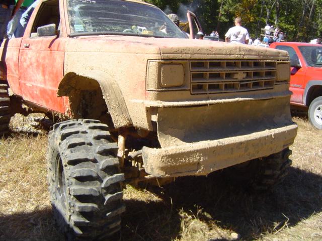 Mud-covered pickup truck with large off-road tires and a custom bumper. Another red pickup truck is visible to the right